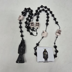 May include: A rosary featuring black faceted beads and four off-white skull beads. A black figure pendant hangs from the bottom. A small square card is included, displaying an illustration of the figure and the text Oraci&oacute;n Contra Da&ntilde;os.