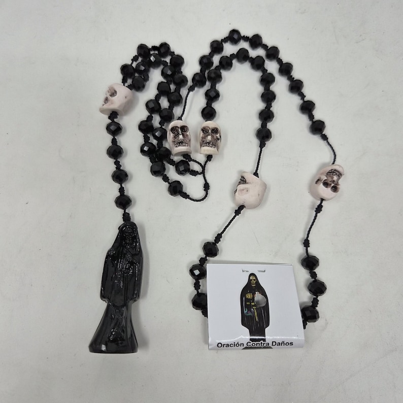 May include: A rosary featuring black faceted beads and four off-white skull beads. A black figure pendant hangs from the bottom. A small square card is included, displaying an illustration of the figure and the text Oraci&oacute;n Contra Da&ntilde;os.