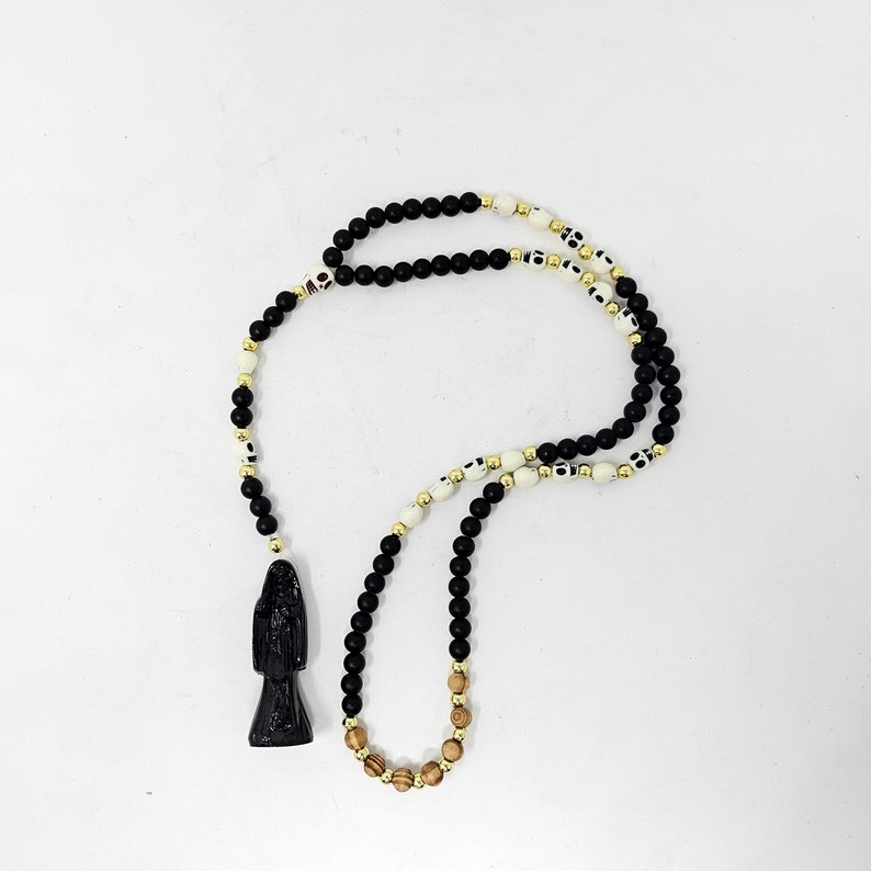 May include: A beaded necklace featuring matte black spherical beads, white skull-shaped beads, small metallic gold spacers, and a few natural wood-toned beads. A black sculpted pendant depicting a robed figure hangs from the end of the strand.