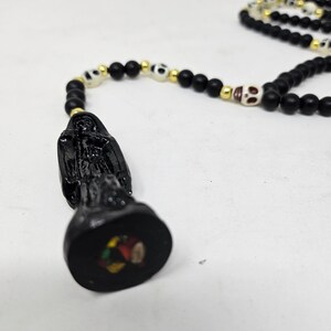 May include: A rosary-style necklace featuring matte black beads, small gold-tone spacers, and off-white skull-shaped beads. The pendant is a black figurine of a hooded figure standing on a circular base with a small, colorful circular inlay.