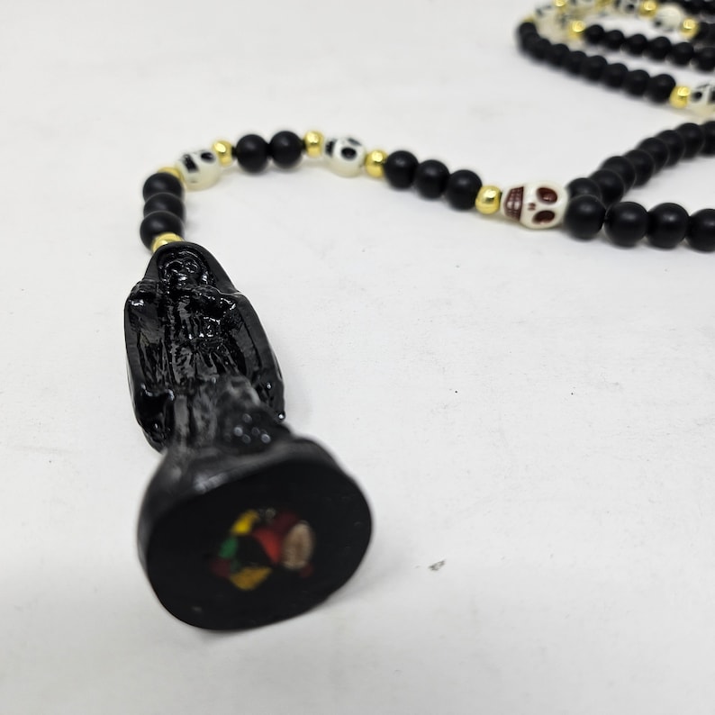 May include: A rosary-style necklace featuring matte black beads, small gold-tone spacers, and off-white skull-shaped beads. The pendant is a black figurine of a hooded figure standing on a circular base with a small, colorful circular inlay.