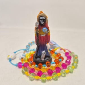 May include: A small figurine of a hooded skeletal figure wearing a red, orange, and purple robe, holding a staff and a blue globe. The figure stands on a black base surrounded by a circular arrangement of colorful translucent beads on string.