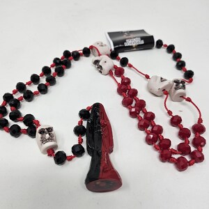 May include: A necklace featuring black and red faceted beads strung on red cord, interspersed with white skull-shaped beads. A central pendant depicts a figure split vertically in black and red. A small tag attached reads ORACION REVERSIBLE.