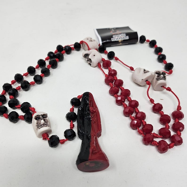 May include: A necklace featuring black and red faceted beads strung on red cord, interspersed with white skull-shaped beads. A central pendant depicts a figure split vertically in black and red. A small tag attached reads ORACION REVERSIBLE.