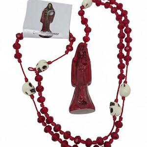 May include: A red rosary featuring faceted red beads and off-white skull-shaped beads. A central red figurine depicts a hooded figure holding a heart. A small white card is attached, displaying an image of the figurine and the text Coraz&oacute;n del Amor.