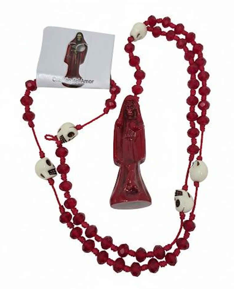 May include: A red rosary featuring faceted red beads and off-white skull-shaped beads. A central red figurine depicts a hooded figure holding a heart. A small white card is attached, displaying an image of the figurine and the text Coraz&oacute;n del Amor.