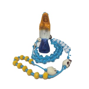 May include: A rosary featuring a small figurine of a hooded figure with an orange head, white torso, and blue base. The rosary is strung on blue cord with faceted light blue beads, opaque yellow beads, white beads, and small white skull-shaped beads.
