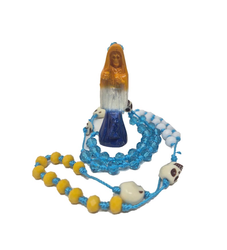 May include: A rosary featuring a small figurine of a hooded figure with an orange head, white torso, and blue base. The rosary is strung on blue cord with faceted light blue beads, opaque yellow beads, white beads, and small white skull-shaped beads.