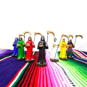 May include: Seven skeletal figures in hooded robes, each holding a scythe and a crystal ball. The figures are painted in different colors, including red, green, blue, black, white, yellow, and purple. They are standing on a colorful striped blanket.