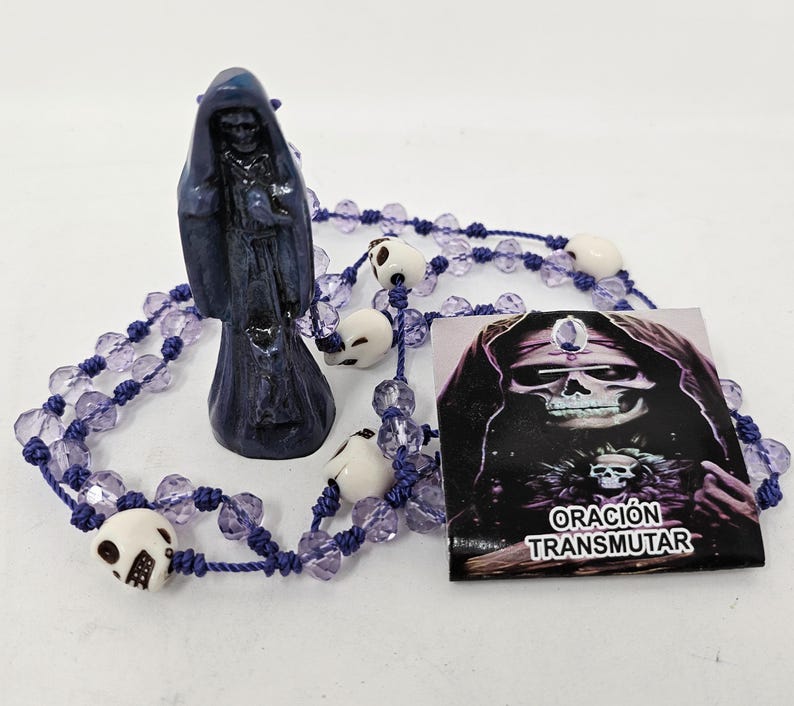 May include: A dark blue hooded figure figurine standing next to a string of purple faceted beads interspersed with small white skull beads. A square card features a skull illustration and the text ORACI&Oacute;N TRANSMUTAR in white capital letters.