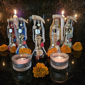 May include: Five Santa Muerte statues in red, blue, black, gold, and silver robes, each holding a scythe and adorned with coins, mirrors, and flowers. Two lit white taper candles stand behind the figures, and two glass tea light holders with lit candles sit in the foreground, surrounded by orange marigold flowers.
