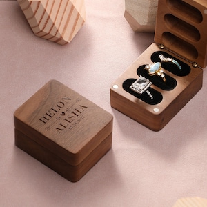 Könnte beinhalten: Eine Holzkiste für Ringe mit einem Scharnierdeckel. Die Kiste ist mit den Namen "Helon & Alisha" und dem Datum "07.21.2022" graviert. Die Kiste ist geöffnet und enthält drei Ringe.