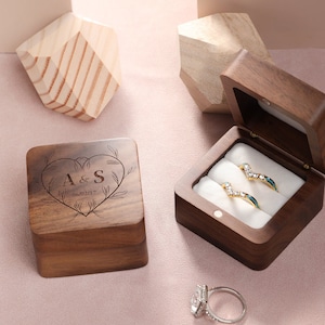 Puede incluir: Dos cajas de anillos de madera. Una caja está cerrada y tiene un corazón con las iniciales "A & S" y la fecha "06/02/17" grabadas en la parte superior. La otra caja está abierta y tiene dos anillos dentro. Los anillos son de plata con pequeños diamantes y una piedra azul.