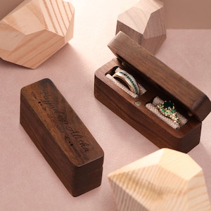 May include: A wooden ring box with a hinged lid, engraved with the text "Holly & Joshua 07.21.2023". The box is open, revealing two rings inside. One ring is a simple band, and the other is a ring with a large green gemstone.