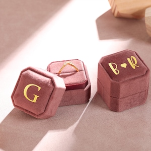 Könnte beinhalten: Zwei rosafarbene Samt-Ringboxen mit goldenen Buchstaben. Eine Box sagt "G" und die andere sagt "B ♥ R". Ein goldener Ring mit kleinen Diamanten befindet sich in einer der Boxen.
