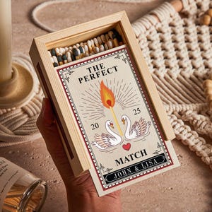 Może przedstawiać: Drewniana skrzynka zapałek z czerwoną i białą etykietą, na której widnieje napis "The Perfect Match" oraz grafika przedstawiająca dwa łabędzie i serce. Etykieta zawiera również imiona "John & Lisa".