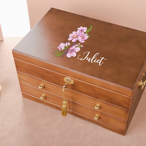 Può includere: Un cofanetto per gioielli in legno con tre cassetti e un design floreale sul coperchio. La scatola è marrone e ha dettagli dorati. Il coperchio è inciso con il nome "Juliet".