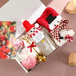 Könnte beinhalten: Eine weihnachtliche Geschenkbox mit einer Grußkarte, einem Schneemann-Plüschtier und anderen festlichen Artikeln. Die Box enthält einen rot-weißen Schneemann-Schal, eine kleine Schneemannfigur, einen herzförmigen Gegenstand und einen goldenen Löffel.