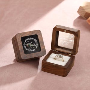 Könnte beinhalten: Zwei Holzkästchen für Ringe mit einem durchsichtigen Glasdeckel. Das Kästchen links hat ein Blumenmuster und den Text "Sarah & Bailey June 17, 2023". Das Kästchen rechts hat einen Ring drin und den Text "Wedding Bells".