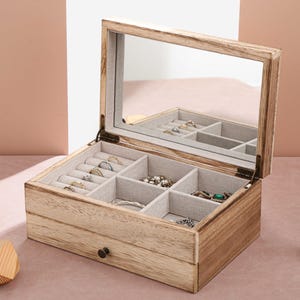 May include: A wooden jewellery box with a mirrored lid and multiple compartments for storing rings, earrings, and other jewellery. The box has a light brown finish and a drawer for additional storage.