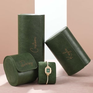 Könnte beinhalten: Vier grüne Samt-Uhrenboxen mit goldenen Beschriftungen. Die Boxen sind mit "Custom", "Daisy" und "Joy" beschriftet. Eine grüne Samt-Uhrenrolle mit einer goldenen Uhr ist ebenfalls abgebildet.