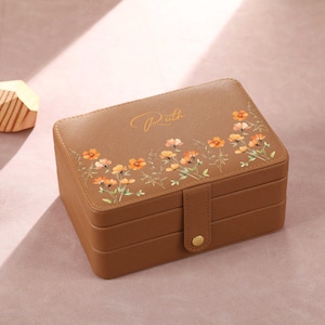May include: A brown leather jewellery box with a floral design and the name "Roth" in script. The box has a snap closure and is stacked with multiple compartments. The floral design features orange and yellow flowers.