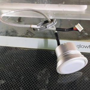 May include: A cylindrical silver metal component with a white circular top, connected to a gray electrical wire featuring a plastic locking ring and a small white multi-pin connector. The wire is marked with the text P1D in black ink.