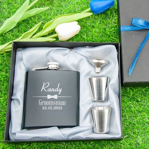 Könnte beinhalten: Eine schwarze Geschenkbox mit einem blauen Band und Schleife. Die Box enthält eine silberne Flachmann mit dem Namen "Randy" und dem Text "Groomsman 22.12.2022" eingraviert. Der Flachmann wird von einem silbernen Trichter und zwei silbernen Schnapsgläsern begleitet.