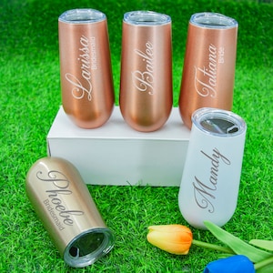 May include: Set of five personalized champagne flutes with names engraved on each. The flutes are in rose gold and white with the words "Larissa Bridesmaid", "Bailee", "Tiana Bride", "Mandy" and "Phoebe Bridesmaid".