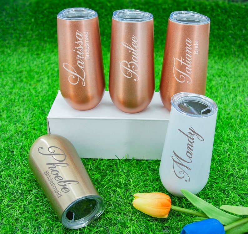 May include: Set of five personalized champagne flutes with names engraved on each. The flutes are in rose gold and white with the words "Larissa Bridesmaid", "Bailee", "Tiana Bride", "Mandy" and "Phoebe Bridesmaid".