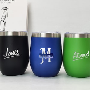 Op de afbeelding: Drie roestvrijstalen wijnglazen met verschillende gepersonaliseerde gravures. Het glas links is zwart met de naam "Jones" gegraveerd. Het glas in het midden is blauw met de naam "Humberto" en de initialen "M" gegraveerd. Het glas rechts is groen met de naam "Atwood" gegraveerd.