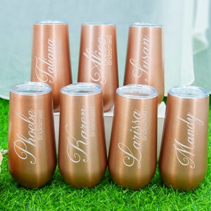 May include: Six rose gold stainless steel tumblers with personalized names and the word "bridesmaid" engraved on each. The tumblers are arranged in a row on a green surface.