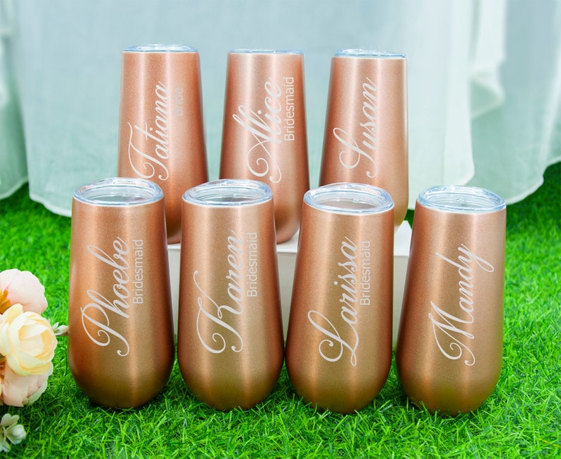 May include: Six rose gold stainless steel tumblers with personalized names and the word "bridesmaid" engraved on each. The tumblers are arranged in a row on a green surface.
