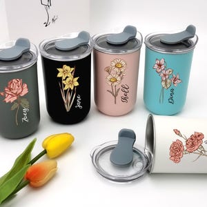 May include: Several insulated tumblers in different colours, each decorated with a floral design and a name. The tumblers have grey lids and are displayed with a tulip. The colours include grey, black, pink, and teal.