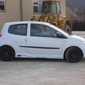 Puede incluir: Un coche blanco compacto con ruedas negras y un borde negro alrededor de las ventanas. El coche est&aacute; aparcado delante de un veh&iacute;culo de construcci&oacute;n amarillo.
