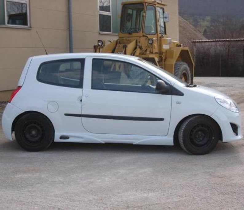 Puede incluir: Un coche blanco compacto con ruedas negras y un borde negro alrededor de las ventanas. El coche est&aacute; aparcado delante de un veh&iacute;culo de construcci&oacute;n amarillo.