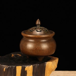 Puede incluir: Un quemador de incienso de bronce con una tapa que presenta un diseño decorativo. El quemador tiene tres patas y un cuerpo redondeado.