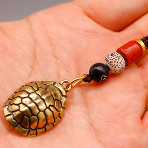 May include: A bronze turtle shell pendant with a gold-coloured bail is attached to a brown cord necklace. The necklace features a series of beads in black, white with brown spots, and red. The pendant is held in a person's hand.