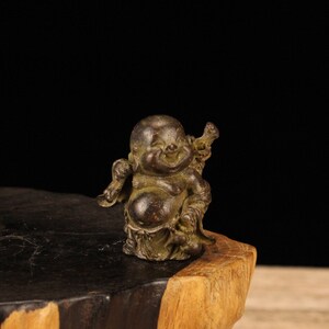 Puede incluir: Una pequeña estatua de bronce de una figura de Buda sonriente y regordeta. La estatua está de pie sobre una base de madera.