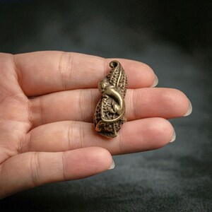 May include: A small, metallic, bronze-colored charm shaped like a peanut with a textured, shell-like surface. A tiny, sculpted lizard or gecko figure is attached to the front of the peanut, featuring a curved tail and detailed body contours.