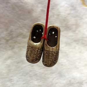 Puede incluir: Un par de zapatos decorativos pequeños de color dorado con diseños intrincados y agujeros. Los zapatos están atados con una cuerda roja.