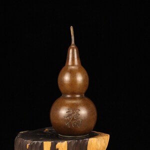 Puede incluir: Un jarrón de metal marrón en forma de doble calabaza con cuello estrecho y una inscripción tallada en la parte inferior. El jarrón tiene un aspecto rústico y antiguo.