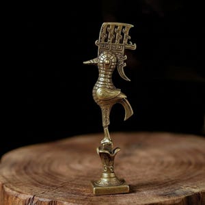 Puede incluir: Una figura de metal color latón de un ave estilizada de pie sobre un pequeño pedestal. El ave presenta una cresta alta e intrincada con patrones geométricos en la cabeza y un diseño texturizado de rejilla que cubre su cuerpo y alas.