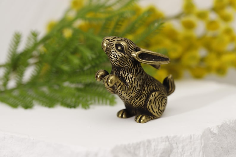 Puede incluir: Una figura de conejo de color bronce en posici&oacute;n de pie. El conejito tiene una textura de pelaje detallada y se encuentra sobre un fondo de follaje verde y amarillo. La figura mide aproximadamente 5 cm de altura.