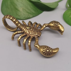 Op de afbeelding: Een gouden metalen scorpion-bedel met ingewikkelde details. De scorpion heeft een gekrulde staart en een gedetailleerd ontwerp op zijn rug.