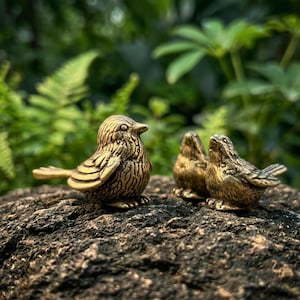 Puede incluir: Tres pequeñas figuras de pájaros de latón con texturas de plumas grabadas detalladas. Los pájaros están representados en posición de reposo con cuerpos redondeados, picos puntiagudos y alas plegadas, con un acabado metálico dorado.