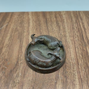 May include: A bronze sculpture of two stylised animals, possibly goats, facing each other in a circular base.