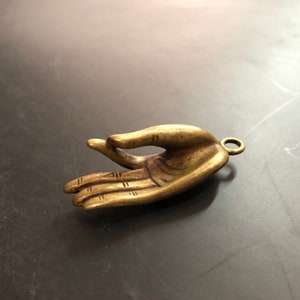 Puede incluir: Un amuleto de mano de latón con un anillo para colgar. La mano está en un gesto de dar o recibir.