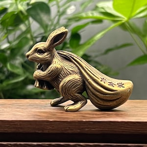 Pode incluir: Uma estatueta de coelho bronzeada carregando um saco grande. O coelho é detalhado com pelo gravado e segura uma pequena bolsa. O saco tem caracteres chineses. A estatueta está em uma superfície de madeira com um fundo verde.