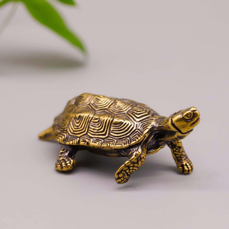 May include: A small, detailed brass turtle figurine. The turtle has a textured shell with intricate patterns and is posed with its legs extended. The figurine is a warm gold color and appears to be made of metal.